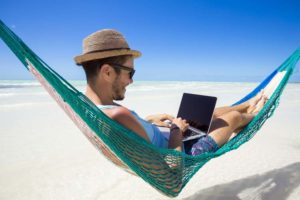 Happy man working remotely with a laptop, on a hammock at the beach.